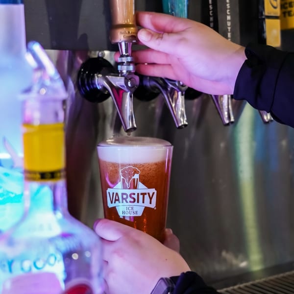 A person's hands filling a Varsity Ice House branded glass with beer from a tap.