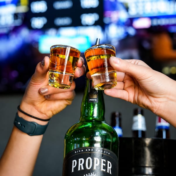 Two hands holding shot glasses of amber liquid over a green bottle of Proper Twelve whiskey.
