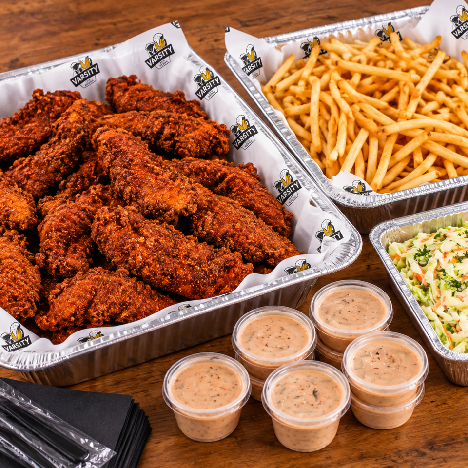 Trays of crispy fried chicken tenders, fries, creamy coleslaw, and dipping sauces on a wooden table.
