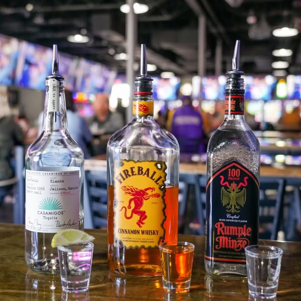 Three liquor bottles with pour spouts and three shot glasses on a reflective bar top.
