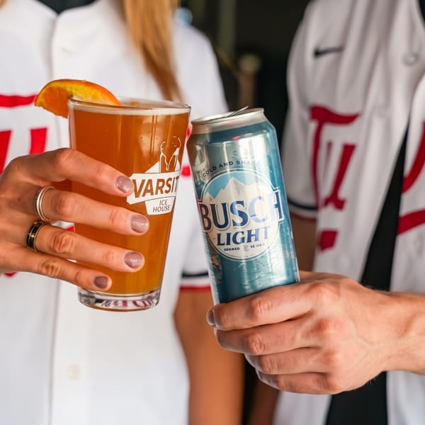 Two people in baseball jerseys clinking a beer glass with an orange slice and a Busch Light can.
