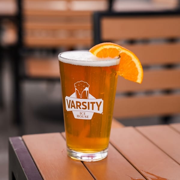 A pint glass of beer with a Varsity Ice House logo and an orange slice on a wooden table.