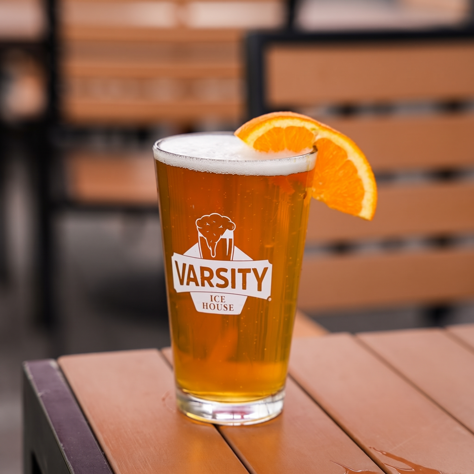 A pint glass of beer with a Varsity Ice House logo and an orange slice on a wooden table.