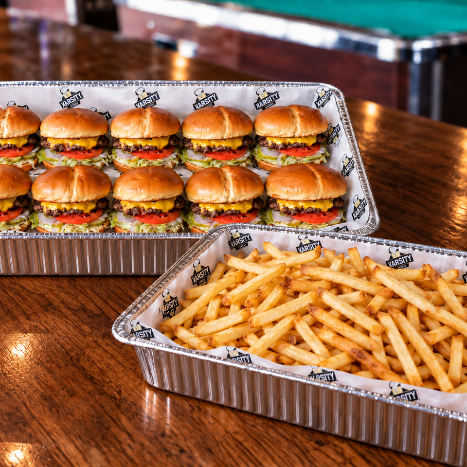 Two foil trays hold ten cheeseburgers with lettuce, tomato, and onions alongside a generous serving of fries on a wooden table.