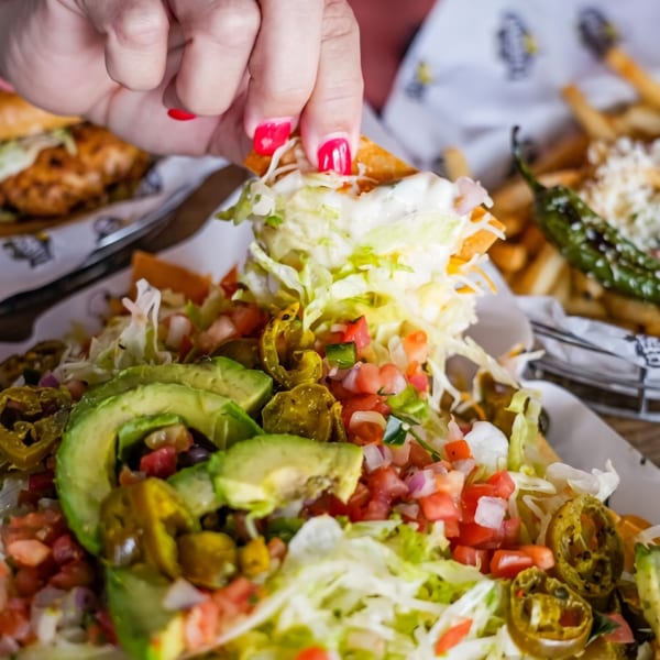 A hand with red nail polish picks up a chip from a plate of loaded nachos with avocado and jalapeños.