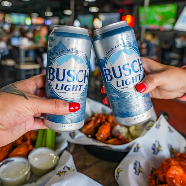 Two hands clinking Busch Light beer cans over baskets of chicken wings and dipping sauces.