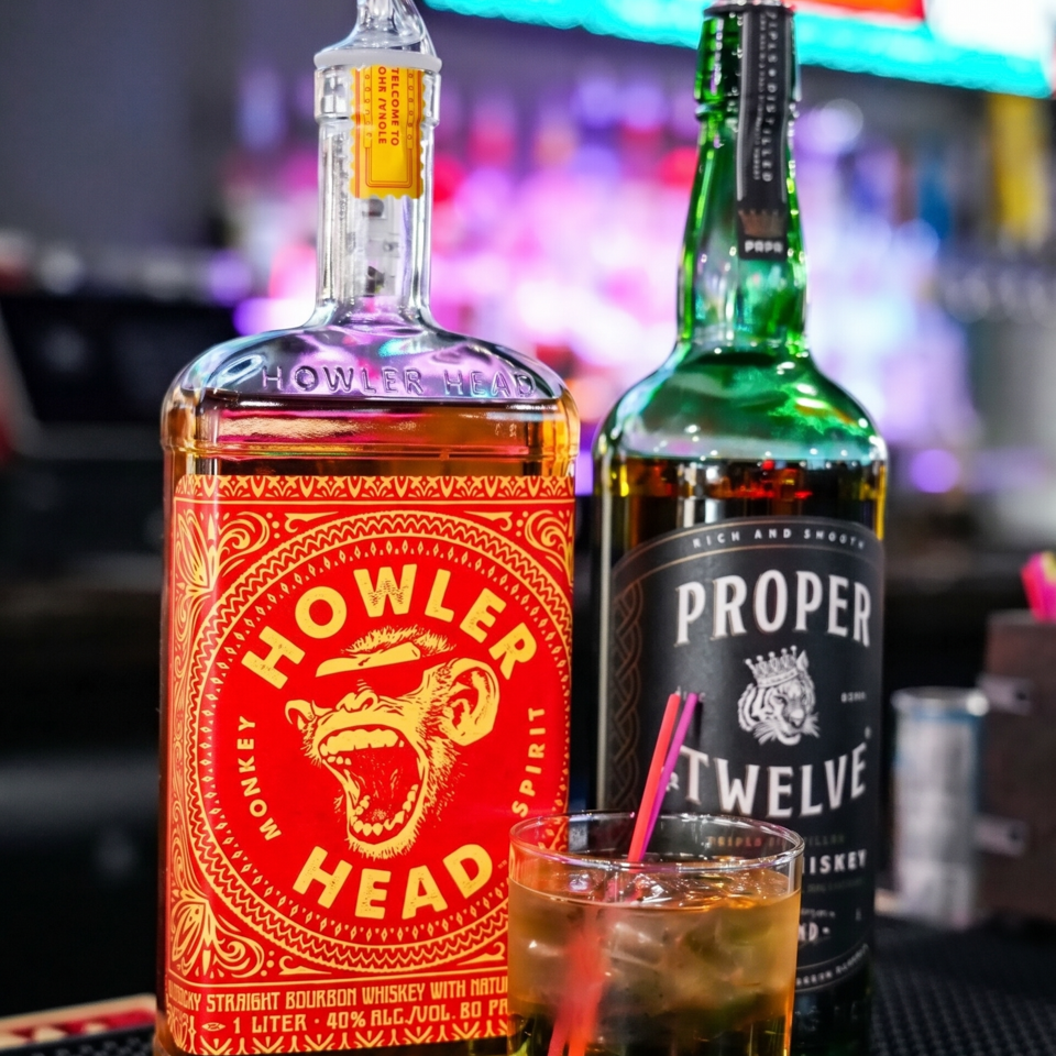 Two whiskey bottles, Howler Head and Proper Twelve, with a glass of whiskey and ice.