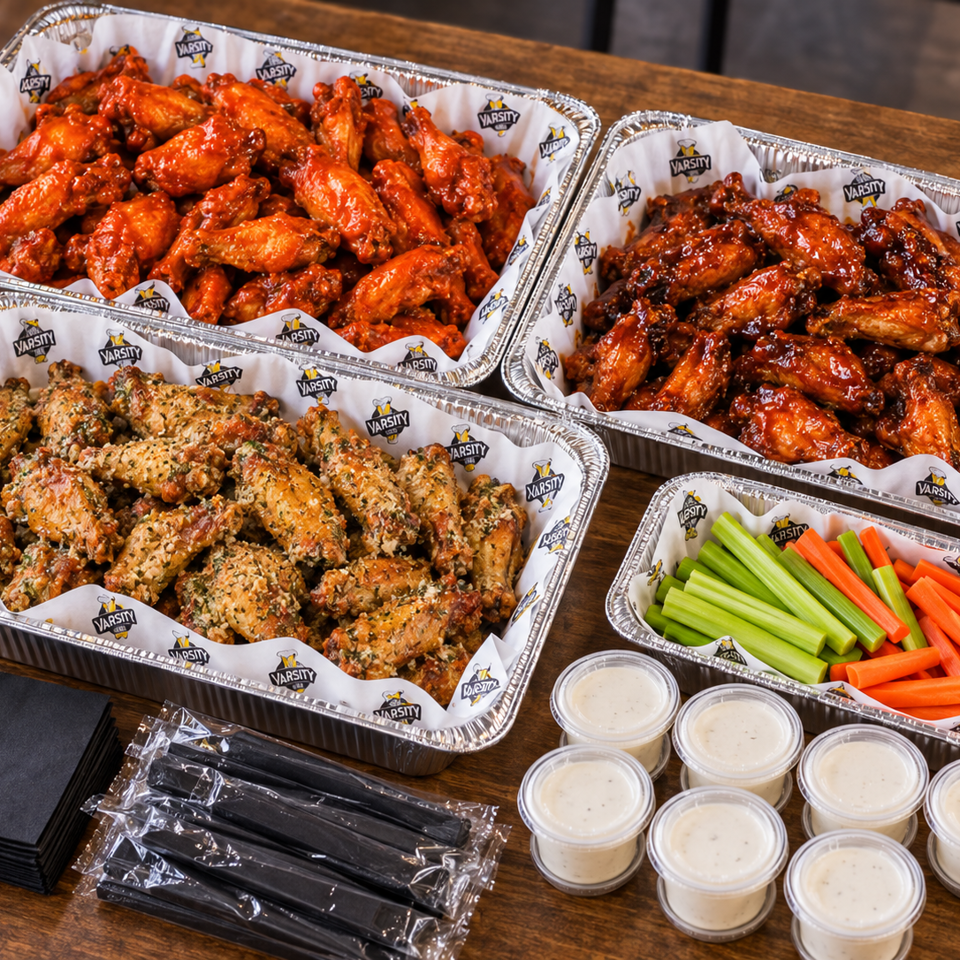 Catering spread of Varsity chicken wings in three flavors, vegetable sticks, dipping sauces, napkins, and utensils on a wooden table.