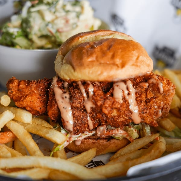 Crispy fried chicken sandwich with sauce, coleslaw, and a side of fries and potato salad.