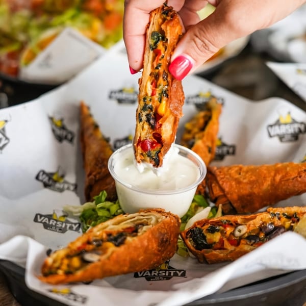 A hand dips a crispy, golden-brown egg roll, filled with colorful vegetables, into a white sauce.