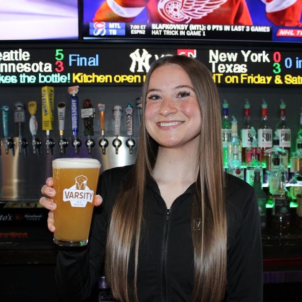 Smiling Varsity Ice House bartender holding a beer, with liquor bottles and sports scores in the background.