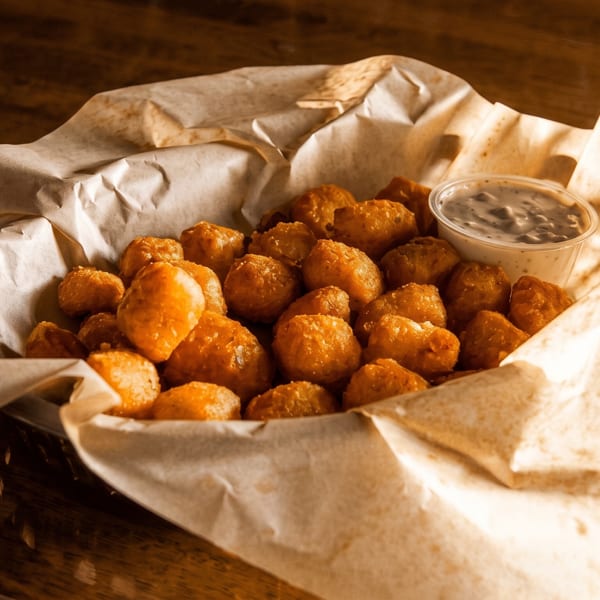 Golden-brown fried cheese curds in a paper-lined basket with a side of dipping sauce on a wooden table.