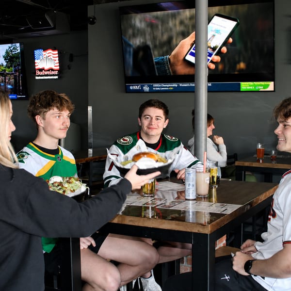 Four young adults enjoying food and drinks at a sports bar with multiple TVs showing games.
