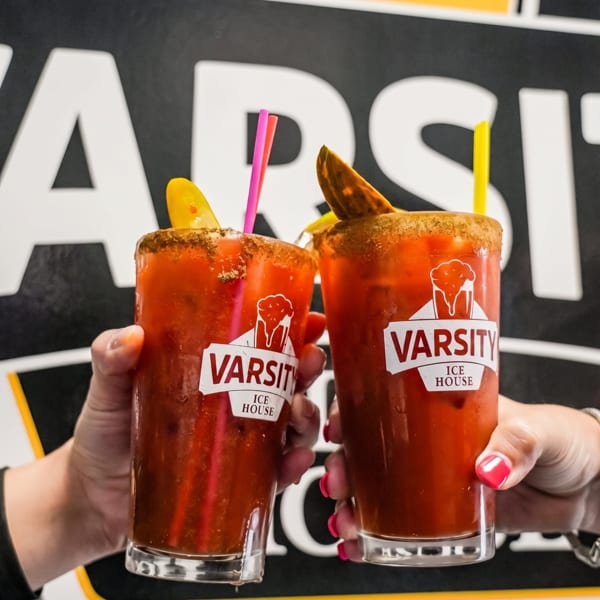 Two hands holding up Varsity Ice House Bloody Mary cocktails with straws and garnishes.